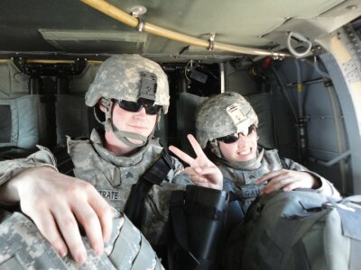 David Westrate in combat uniform gestures happily to the camera alongside another soldier in combat uniform while they both ride in an armored vehicle and leave Iraq.