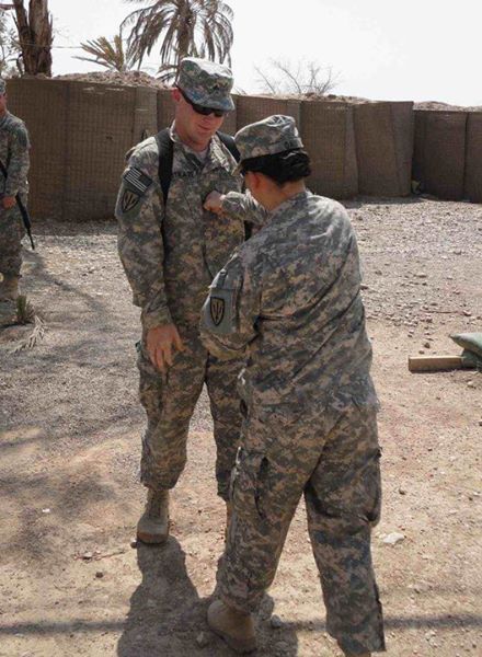 David Westrate has a new rank of Sergeant assigned by another soldier in uniform in a dusty scene inside a large walled compound outdoors.