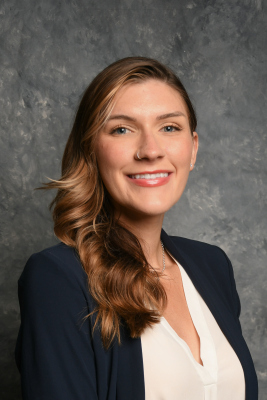 Headshot photo of Amber Renee Quinlan smiling at the camera. She has light brown hair, light skin, blue eyes, and she is wearing a dark blue blazer while standing in front of a swirling gray background.
