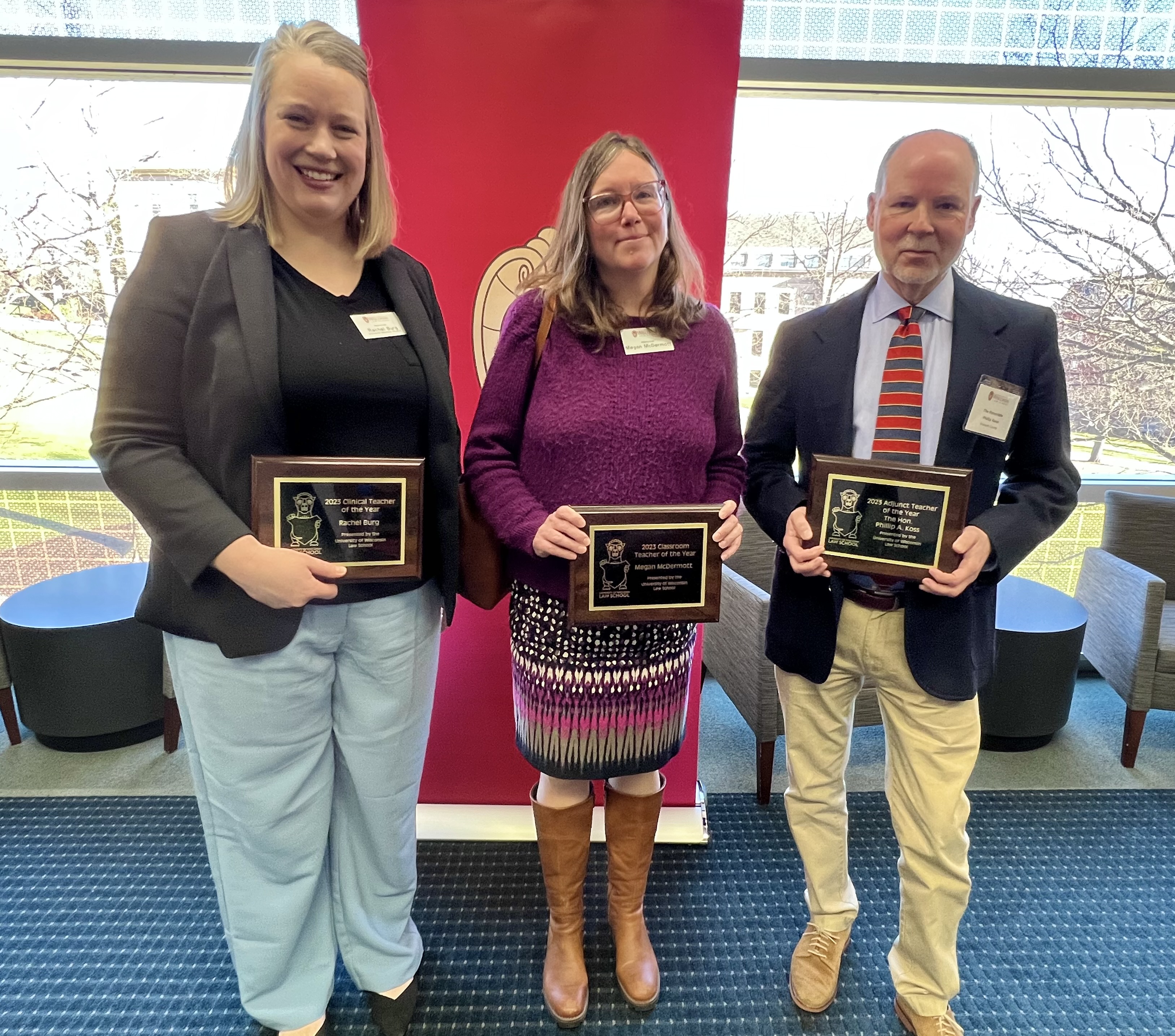 Three people stand together holding plaques for winning the UW Law Teacher of the Year Award. The first, starting from the left, is a tall, blonde woman wearing black with blue slacks and black shoes; the middle is an average height woman wearing purple and a patterned skirt with tall brown boots; and the third is a middle-aged man wearing a dark suit jacket over a light shirt, striped tie, and beige slacks and shoes.