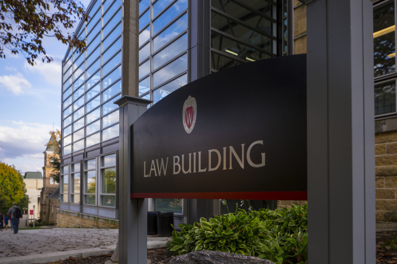 An exterior photo of the Law School building, featuring long horizontal stacked windows and an arched roof against a blue sky. The Law Building sign is out front.