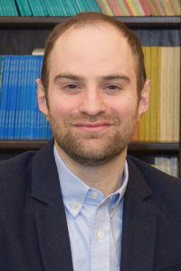 A headshot photo of Ari Schriber smiling with a short cropped beard and wearing a suit jacket while standing in front of a shelf of books