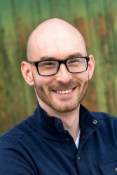 A headshot photo of David Korostyshevsky, who is bald with a thin beard, smiling, and wearing thick black frame glasses and a blue sweater with a zipper