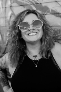 A black and white photo of Doris Morgan Rueda who is smiling with short, wavy styled hair and large, round glasses; their right arm is tattooed