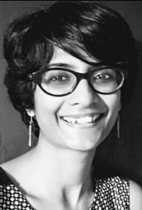 A headshot photo in black and white of Saumyashree Ghosh smiling at the camera with short cut dark hair and wearing thick frame glasses and earrings
