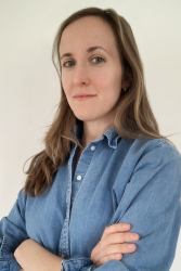 A headshot photo of Linda Kinstler with long blonde hair and standing with arms crossed while wearing a denim shirt