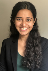 A headshot photo of Maham Theisen smiling with long and curly brown hair while wearing a dark jacket over a green top