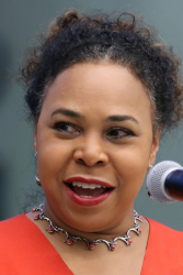 A photo of Michelle McKinley with curly brown hair and wearing a bright colored top while speaking on stage with a microphone nearby