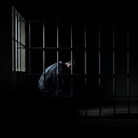 A dark photo-like image of a man sitting solemnly behind bars in the darkness.