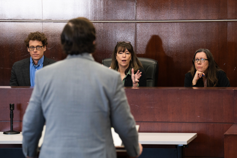 A photo of the back of the young man in a gray suit as he speaks to a panel of judges who appear to be professors.