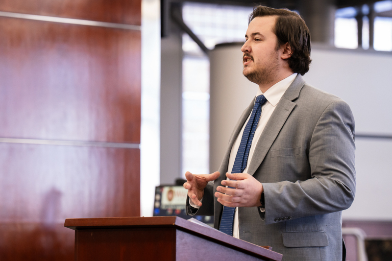 A photo of a young man in a gray suit speaking.