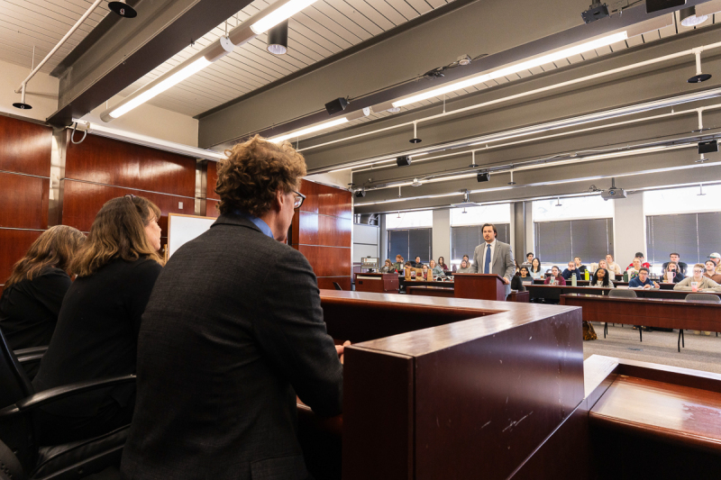 A photo of the room in which a panel of judges, who appear to be professors, sit facing a large group of students.