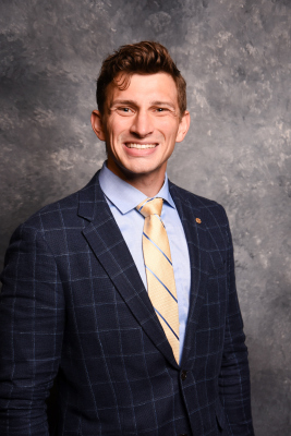 Headshot photo of Treis D. Gunther smiling while wearing a suit in front of a gray background.