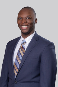 Headshot photo of Brandon Pope - a black man smiling warmly at the camera and wearing a navy blue suit jacket over a white shirt with a gold and sky blue striped tie with a silky texture.