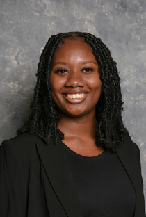 Headshot photo for Ja'Lia Butler, a black woman wearing a black top with a black blazer draped over her shoulders while smiling at the camera in front of a swirly silver and gray background.