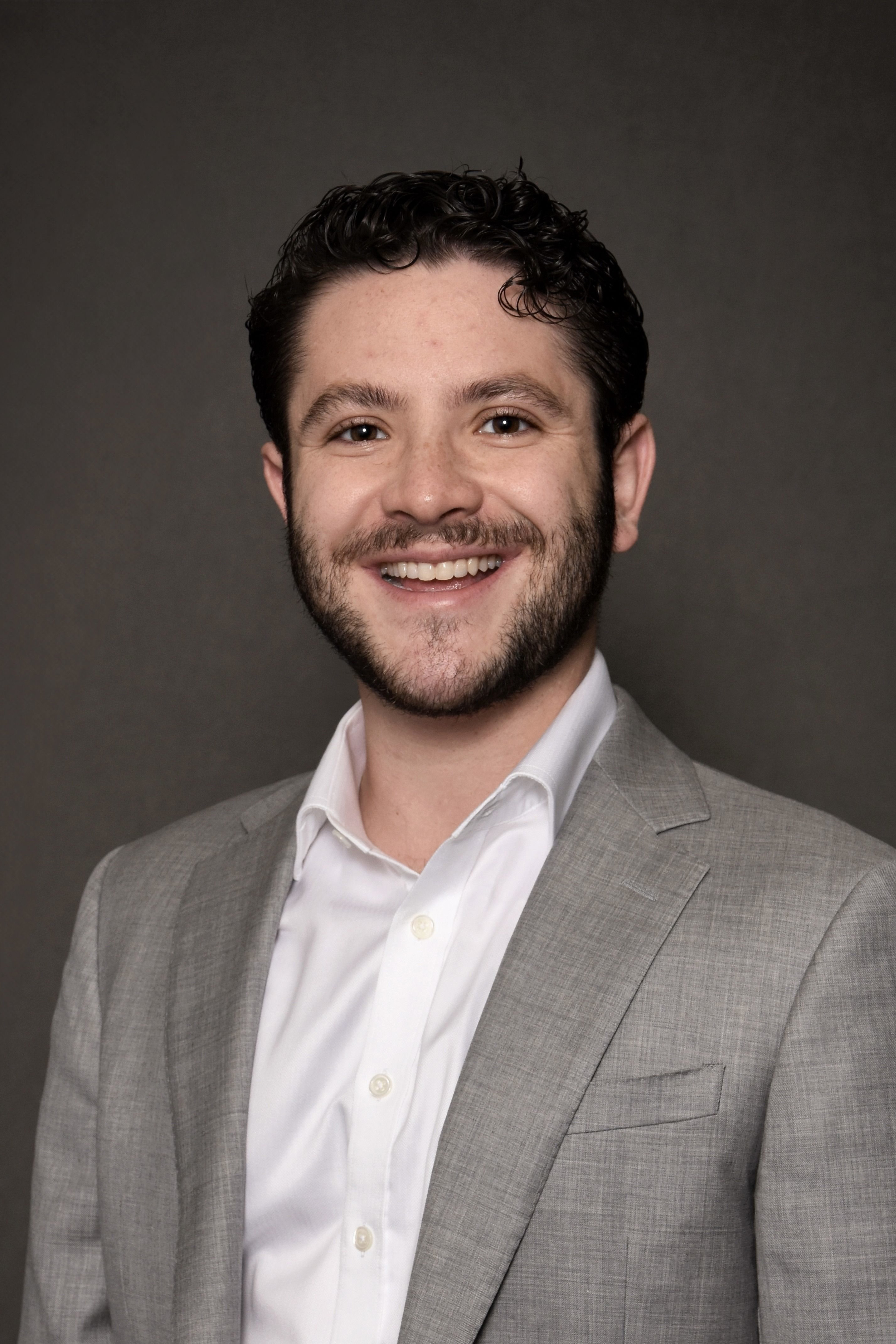 Jake Yalowitz headshot in a grey blazer, white button down shirt and a dark background