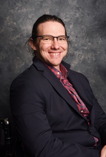 Headshot of Steven Slack smiling at the camera while wearing black partial-frame glasses, a black blazer, and a checkered red shirt.