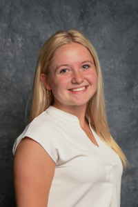 A headshot photo of Gracie Wendt, a blonde haired and fair skinned woman with hazel eyes. She is smiling, wearing a white top, and standing in front of a gray swirled background.