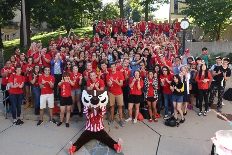 A large group of students stand with Bucky Badger, all making the W hand gesture.